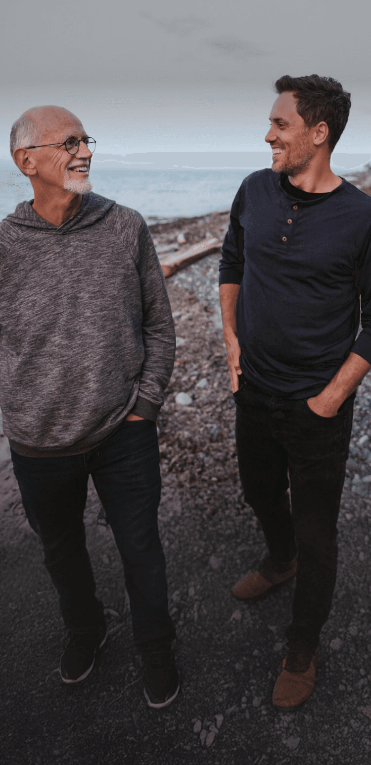 Two men walking and smiling at each other on a rocky beach path, with the ocean in the background.